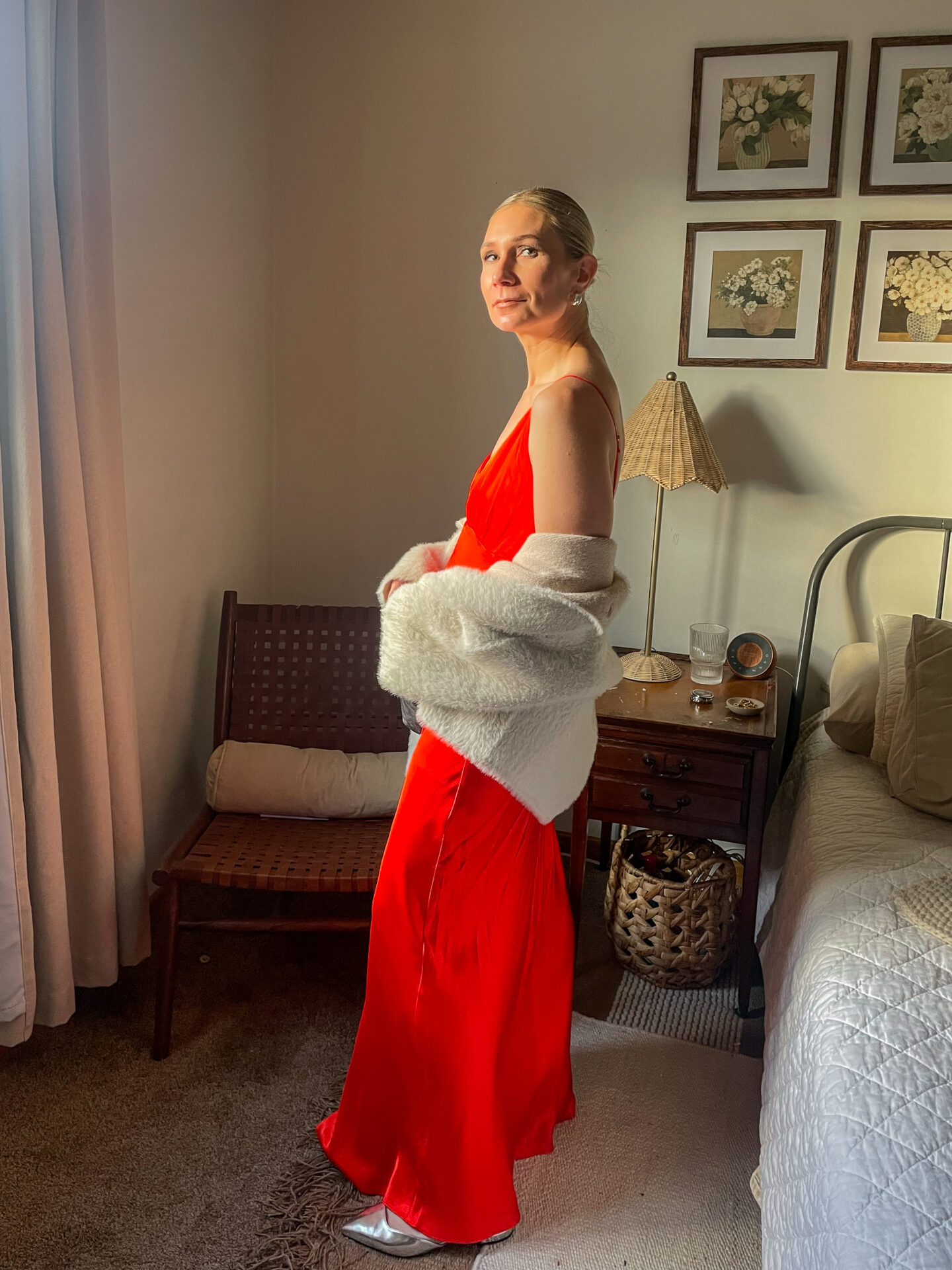 Karin Emily stands in her room wearing a bright red maxi slip dress with a pair of silver shoes and a faux fur drape coat