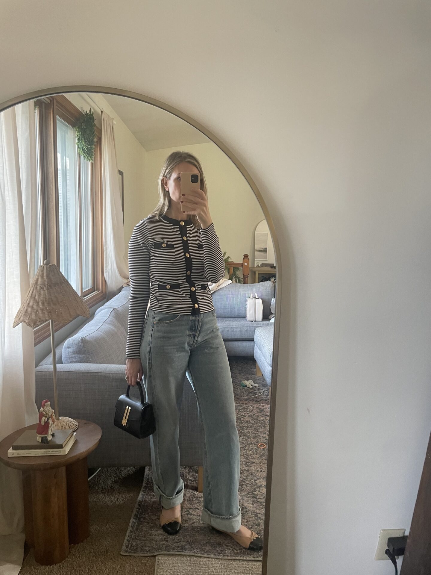 Karin Emily takes a mirror selfie in her living room wearing a striped button down jersey tee with gold buttons and baggy, cuffed jeans with black and nude two tone flats