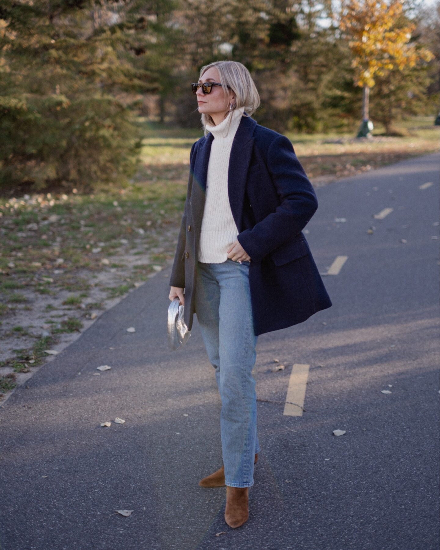 Karin Emily walks down a path wearing a cream turtleneck sweater with a navy wool coat with light wash jeans and brown heeled boots