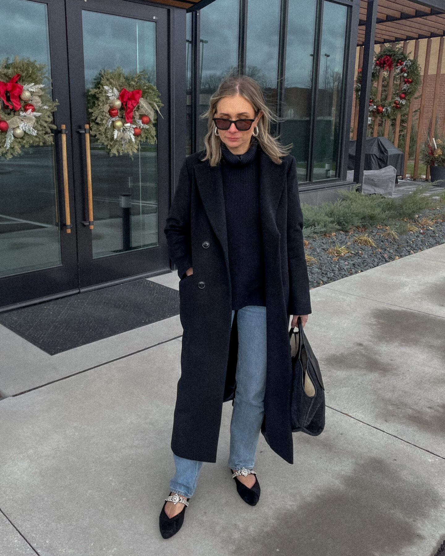 Karin Emily stands in front of a double set of doors with wreaths on them on a sidewalk wearing a long black wool coat over an oversized black turtleneck sweater with a pair of full length light wash jeans and a pair of black flats with a sparkly crystal studded strap