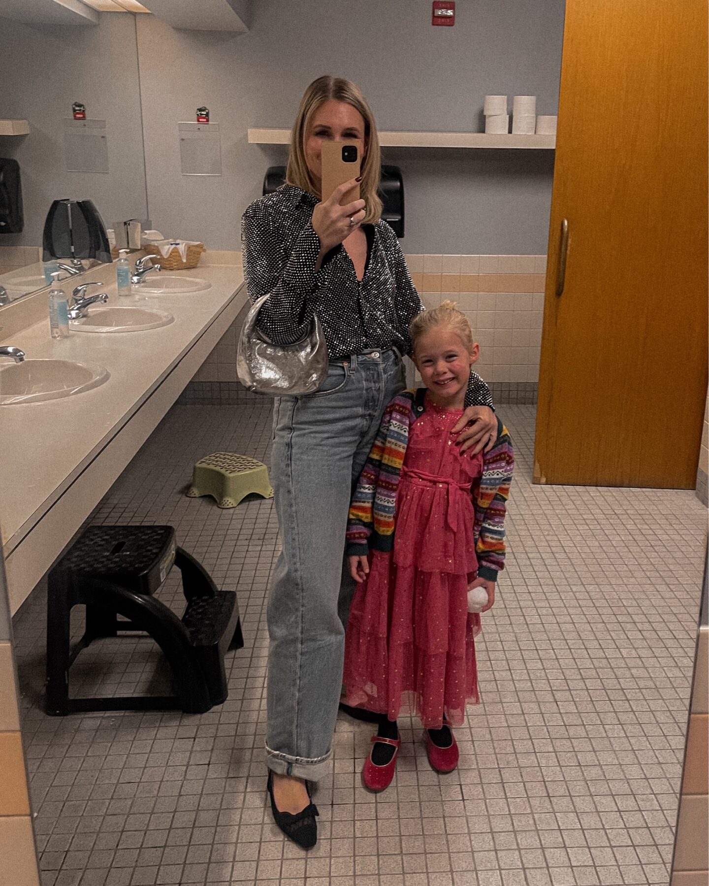 Karin Emily wears a sparkly outfit with a gem studded button down, wide leg ayla jeans from Citizens of Humanity, Black sling back flats and a silver handbag with her daughter who has on a barbie pink sparkly dress with a fair isle rainbow cardigan and rhinestone studded mary jane flats while taking a mirror selfie in a bathroom mirror