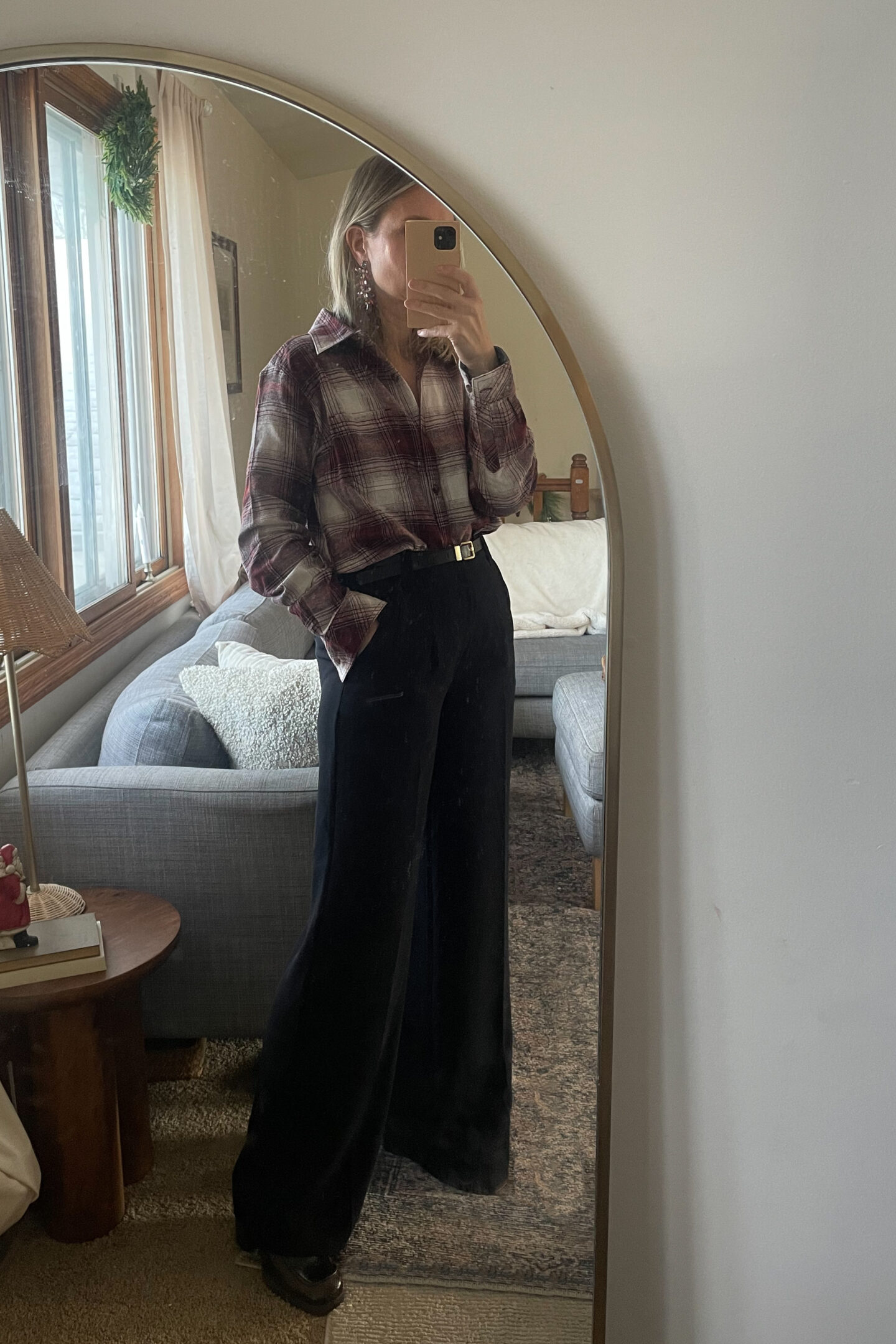 Karin Emily takes a mirror selfie in her living room of a Christmas outfit that includes a burgundy plaid button down top, wide leg black pants, platform loafers, and a pair of sparkly jeweled statement earrings 