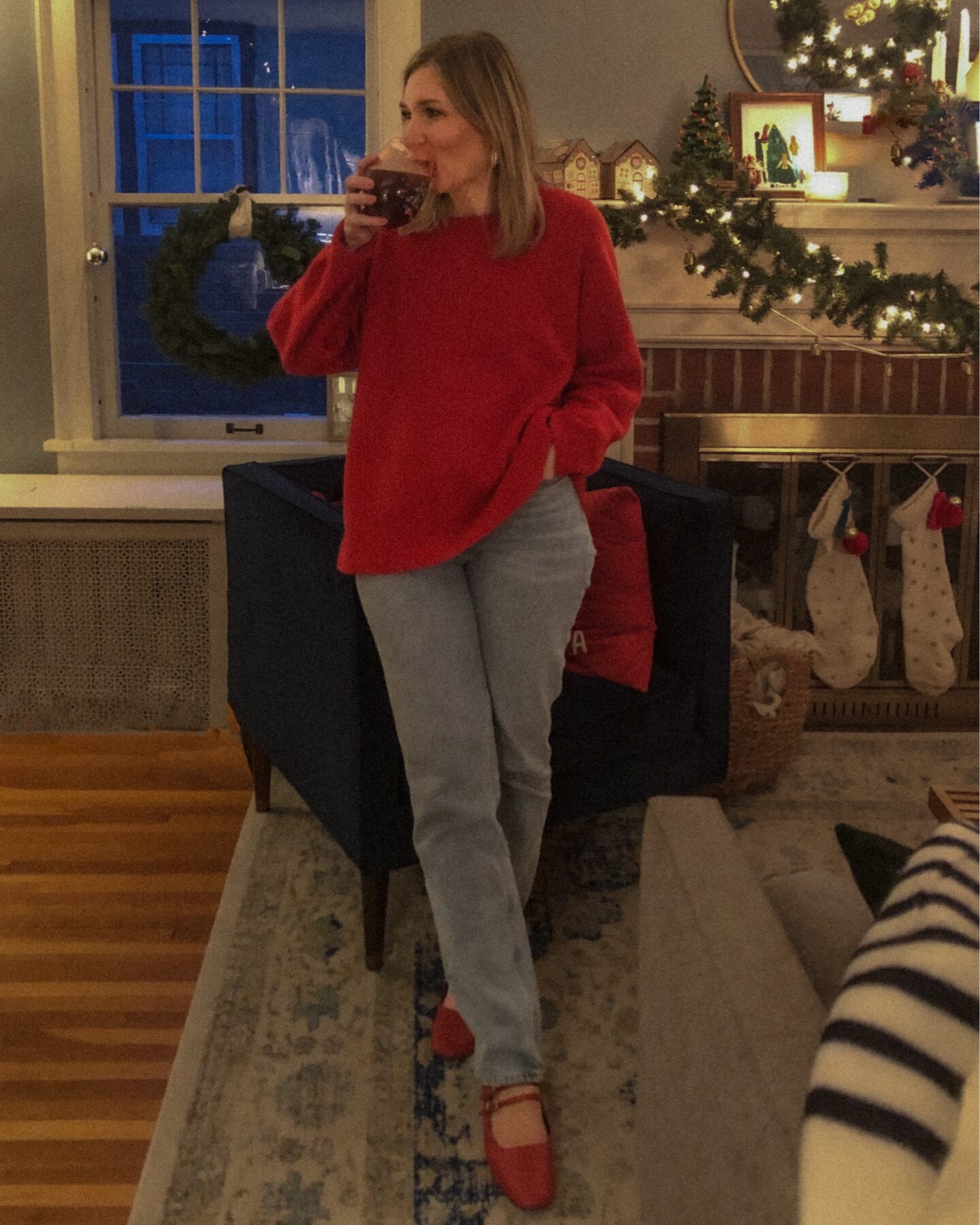 Karin Emily attends a party and stands in a dark living room in front of a navy blue velvet chair and mantel place wearing a red cashmere oversized sweater, light wash jeans and a pair of red Mary Jane flats