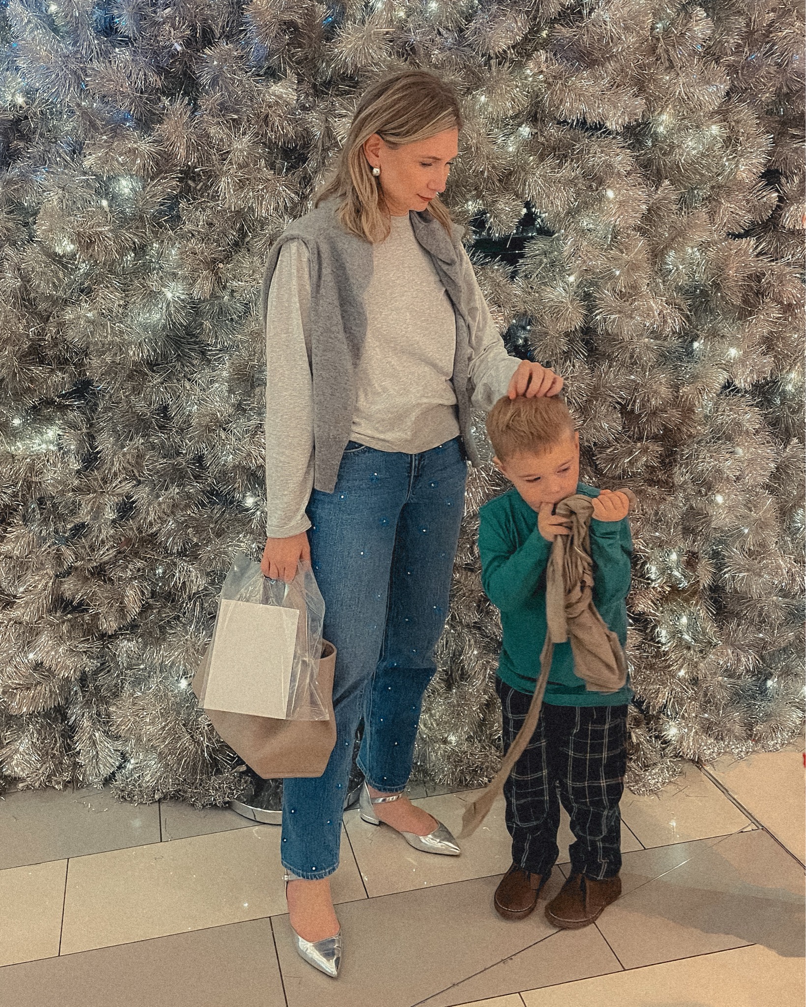 Karin Emily and her son stand in front of a huge silver tinsel tree white white lights and she wears a gray tee with a gray cashmere sweater over her shoulders paired with a pair of sparkly rhinestone studded jeans and silver flats while he wears a green t-rex wearing a Christmas sweater graphic tee over plaid Christmas pants