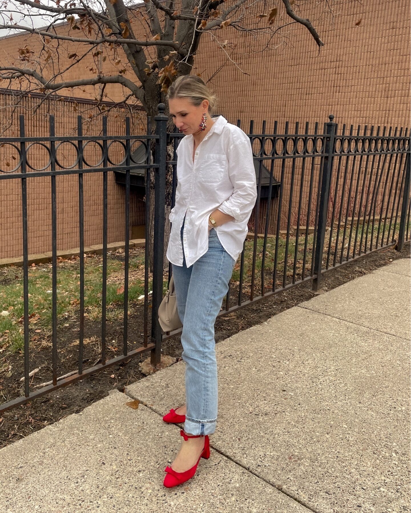 Karin Emily stands on a sidewalk in front of a wrought iron gate wearing an oversized and relaxed white button down with a pair of folded up light wash jeans, a pair of statement and sparkly jeweled earrings, and a pair of red heels with bows on them