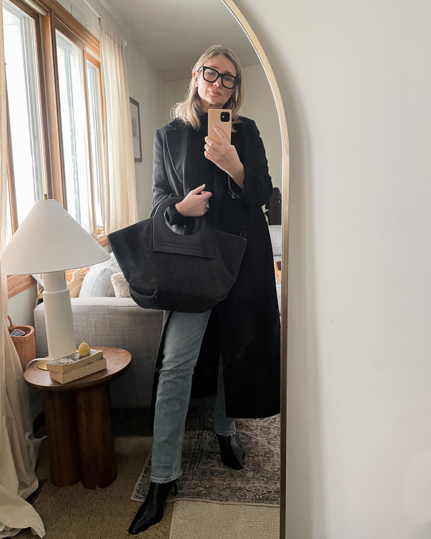 Karin Emily personal style wears a black long wool coat over a black long turtleneck sweater with a pair of agolde light wash jeans and a pair of pointed toe black heeled booties with a hereu black bag