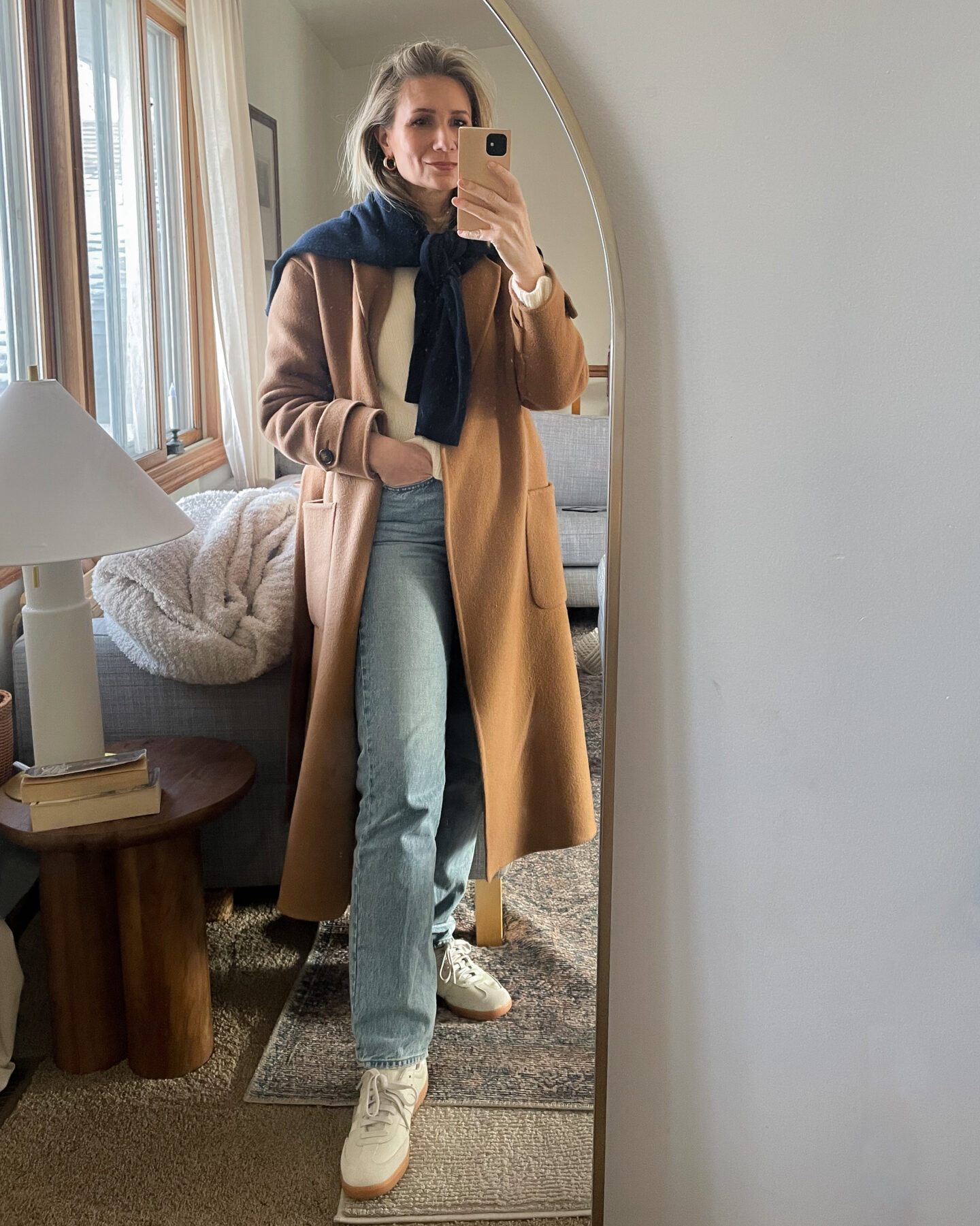 Karen Emily wears a cream cashmere sweater and a pair of light wash Lana jeans from agolde with a cream pair of Adidas Samba sneakers topped with a caramel colored wool wrap coat and a navy cashmere sweater tied like a scarf