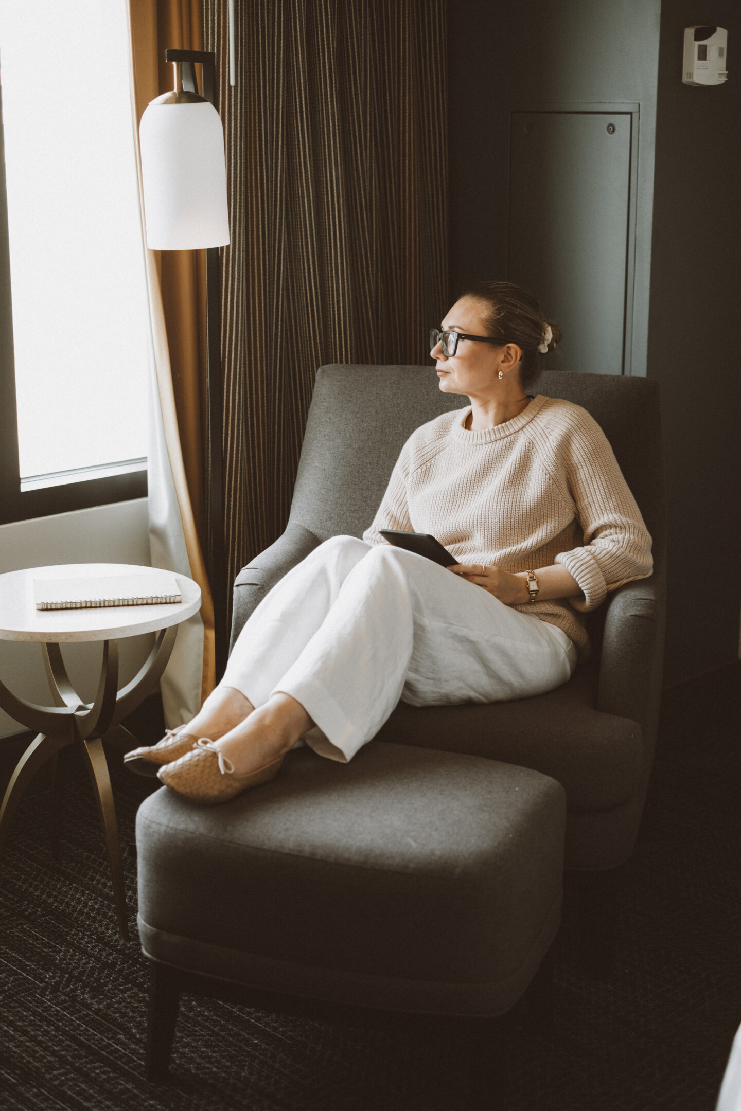 Karin Emily sits in a chair by the window reading her kindle in a oversized sweater, white linen baggy pants, and woven flats from Freda Salvador