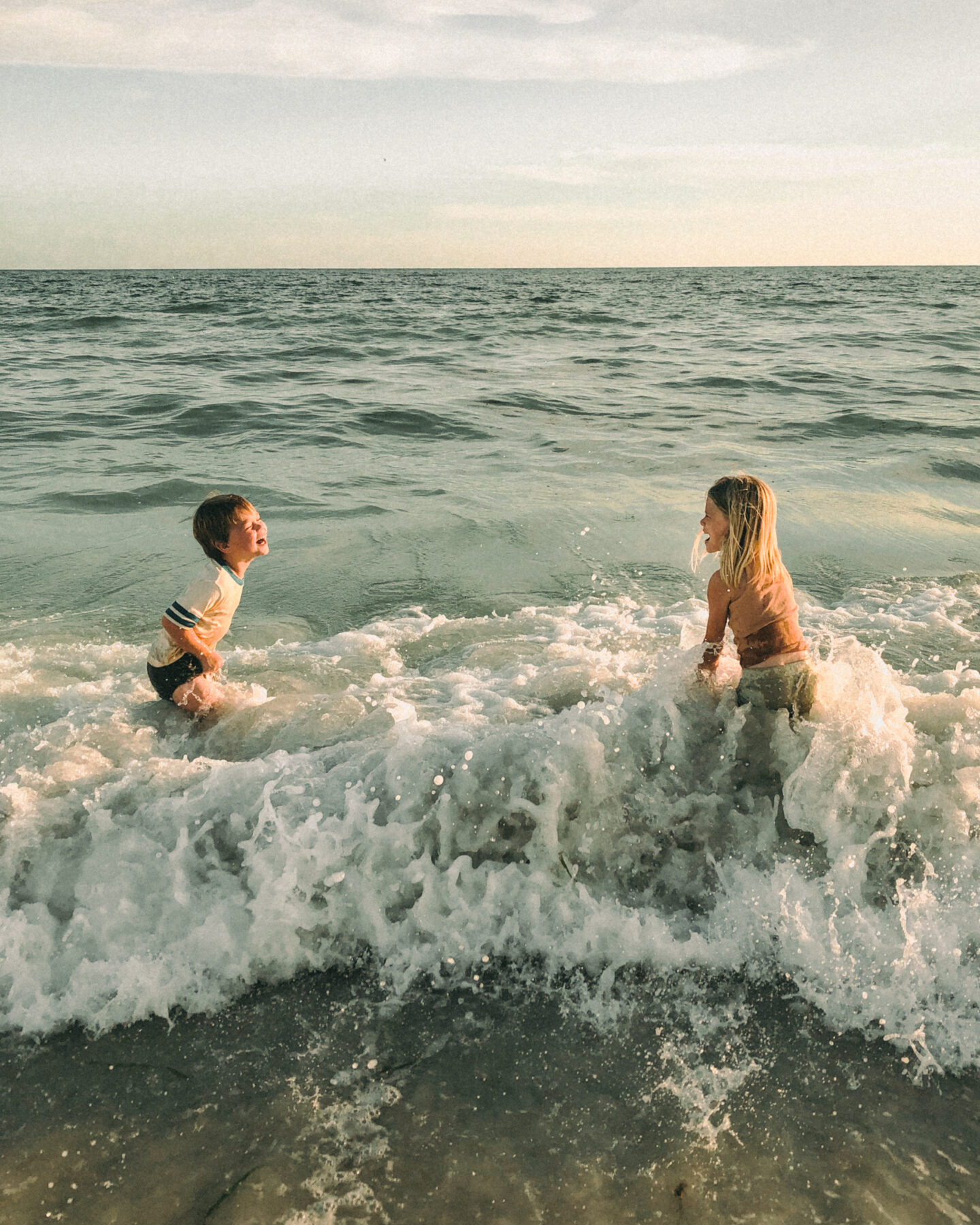 Two young children, a boy and a girl play in the ocean as a wave crashes over them and they laugh with delight. in this blog post Karin Emily talks about having a hysterectomy and what life is like with four neurodivergent children.