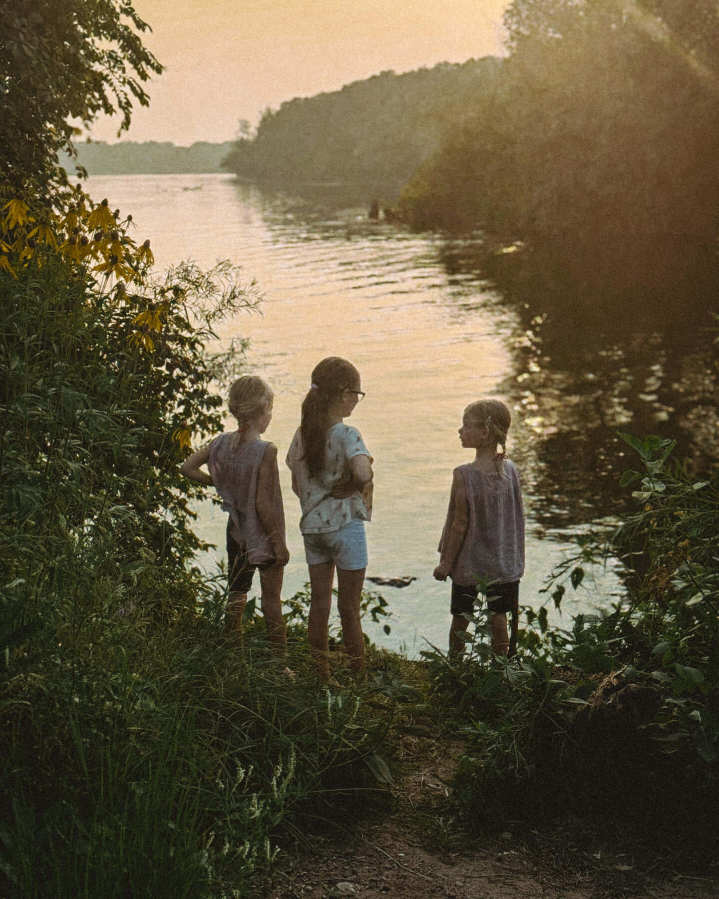 Three young girls stand in front of a river on a grassy embankment at sunset.